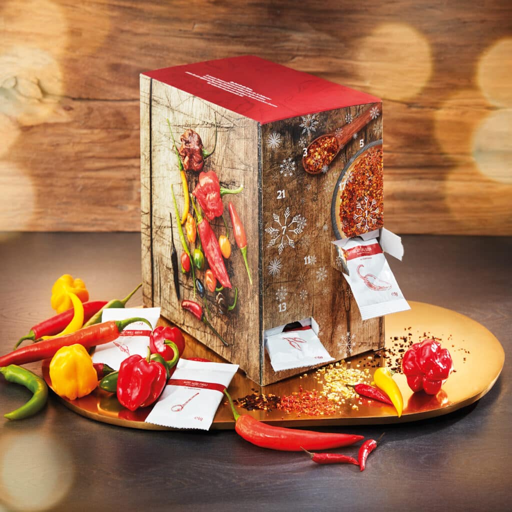Gewürz-Adventskalender mit Chili-Motiven auf Holzhintergrund, umgeben von frischen Chilischoten.