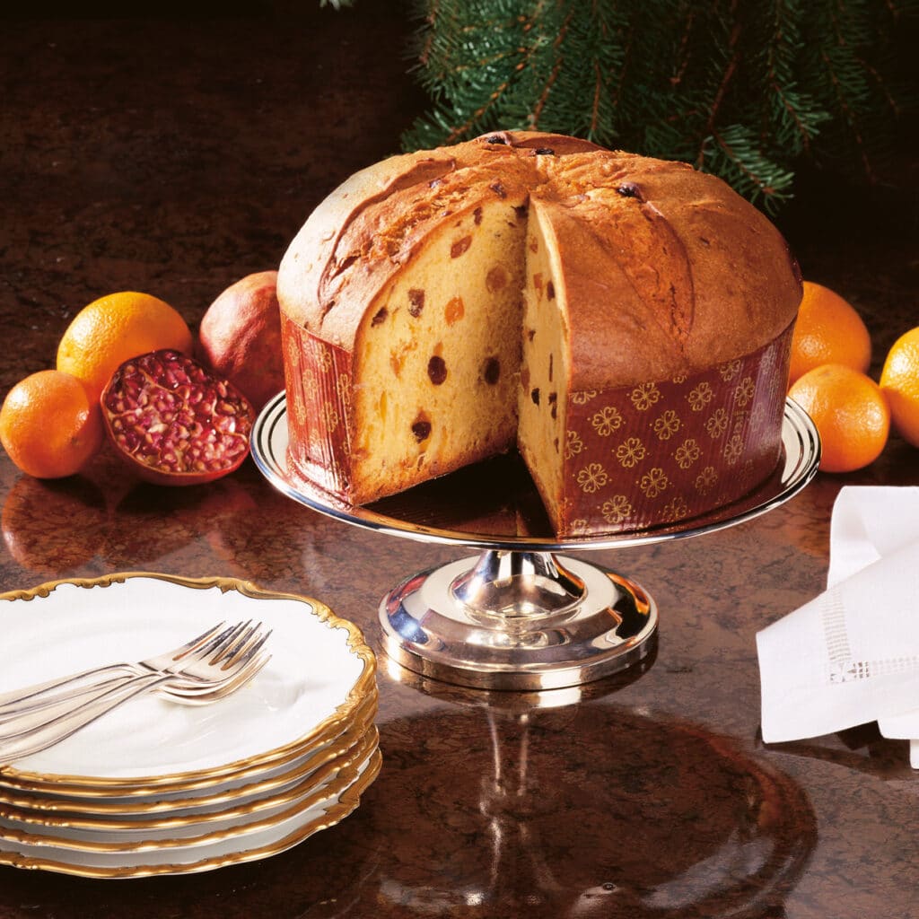 Großer Panettone mit Rosinen auf Kuchenplatte, angeschnitten, umgeben von Früchten und Porzellantellern.