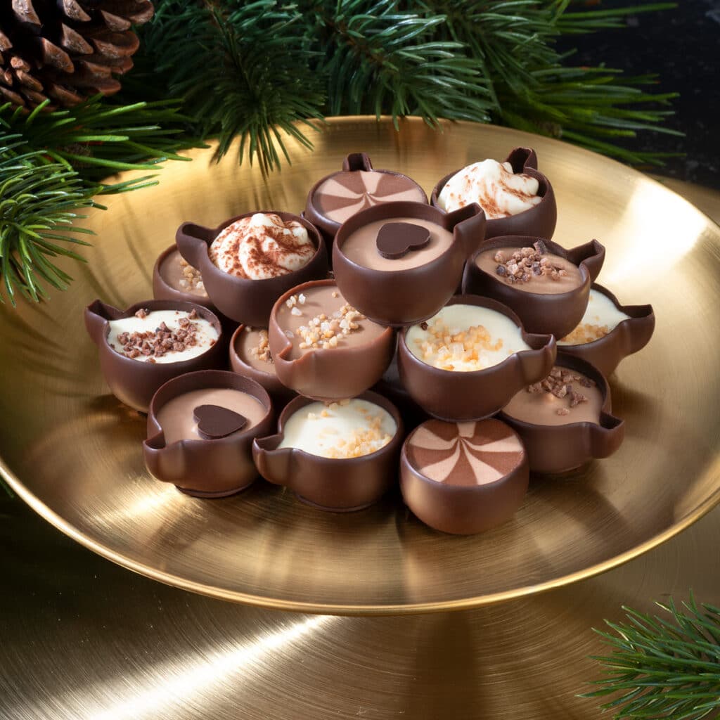 Feine Schokoladenpralinen in kleinen Tassenformen, gefüllt mit verschiedenen Cremes und Nüssen, angerichtet auf einer goldenen Platte mit Tannenzweigen.