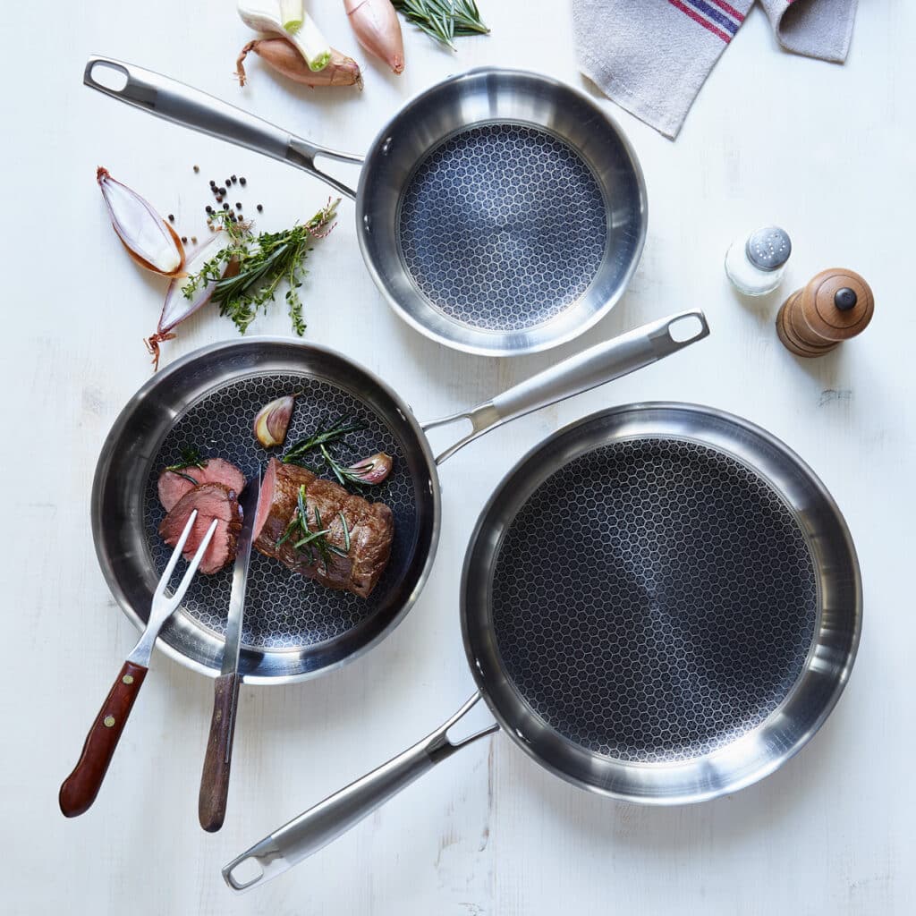 Drei Edelstahlpfannen mit Wabenstruktur, eine mit gebratenem Steak und Kräutern, umgeben von Schalotten.