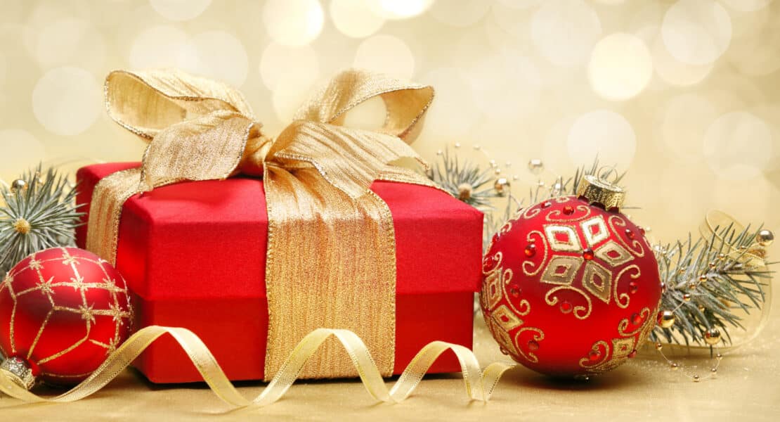 Rotes Weihnachtsgeschenk mit goldener Schleife und glitzernden Christbaumkugeln vor goldenem Bokeh-Hintergrund.