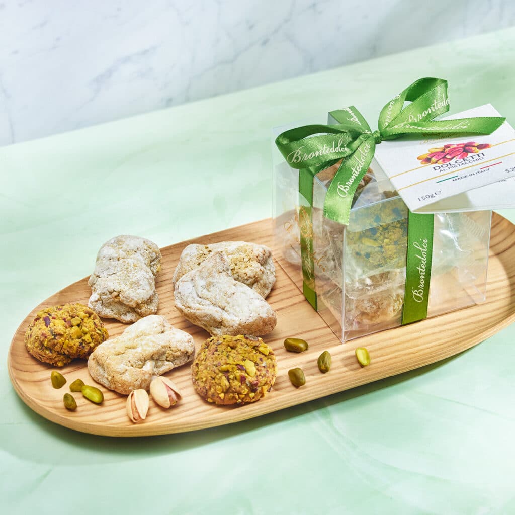 Sizilianische Dolcetti in Geschenkbox mit grüner Schleife, Gebäckvielfalt aus Mandeln und Pistazien auf Holzbrett.