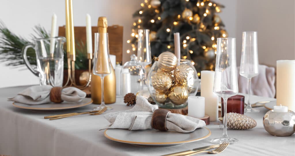 Eleganter Weihnachtstisch mit grauer Tischdecke, goldenen Kerzenständern, Gläsern und weihnachtlicher Deko vor geschmücktem Baum.