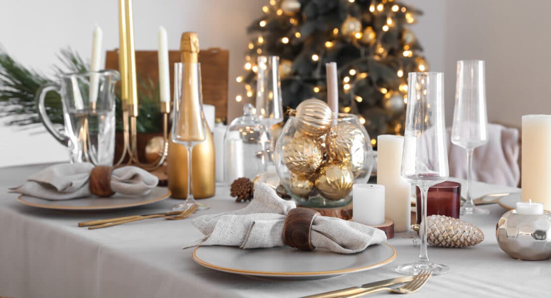 Eleganter Weihnachtstisch mit grauer Tischdecke, goldenen Kerzenständern, Gläsern und weihnachtlicher Deko vor geschmücktem Baum.