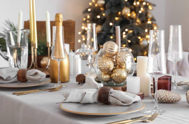 Eleganter Weihnachtstisch mit grauer Tischdecke, goldenen Kerzenständern, Gläsern und weihnachtlicher Deko vor geschmücktem Baum.