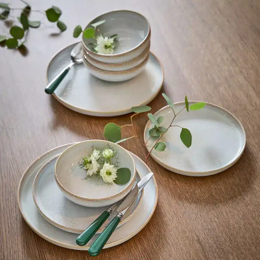 Stapel aus Tellern und Schalen aus hellem Keramikgeschirr mit grünem Besteck und Zweigen auf Holztisch