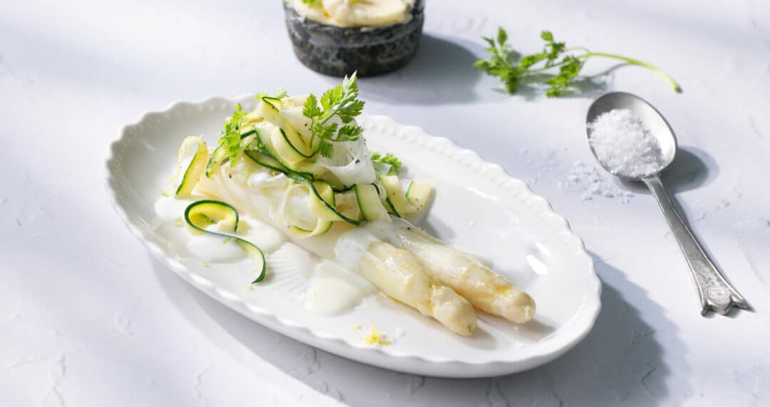 Wei&szlig;er Spargel auf ovalem Teller mit Zucchinistreifen, Kr&auml;utern und Zitronenzeste, Salzl&ouml;ffel daneben